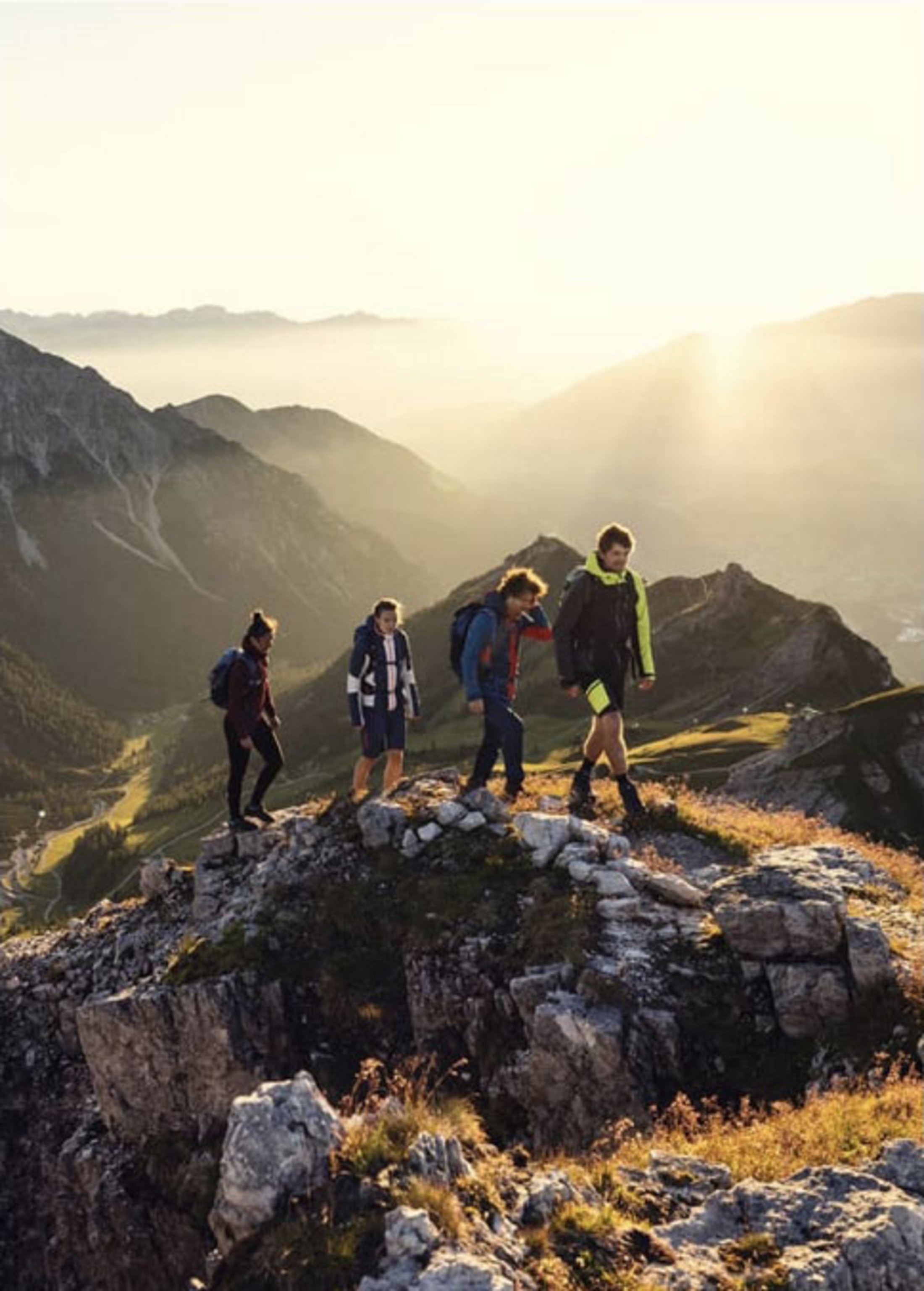 Gruppe von vier Leuten wandern auf einem Berg, im Hintergrund sieht man viele Berggipfel und Sonnenstrahlen.